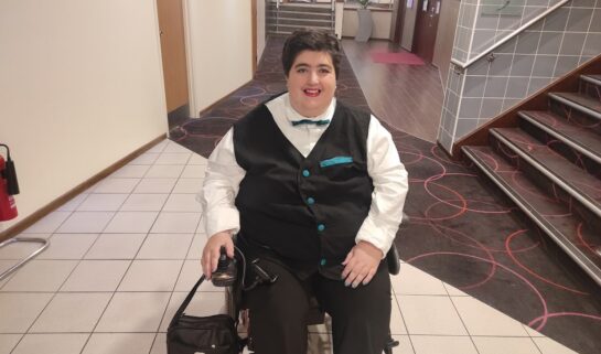 A full-length photo of Morgan smiling in a brightly lit hallway. They are sitting in a power wheelchair, dressed formally in a white collared shirt, a black waistcoat with teal buttons and a teal pocket square, a teal bowtie, and black trousers. A small black bag is attached to the side of the wheelchair. The background shows a hotel-style corridor with neutral-coloured walls, a patterned carpet, and a staircase.