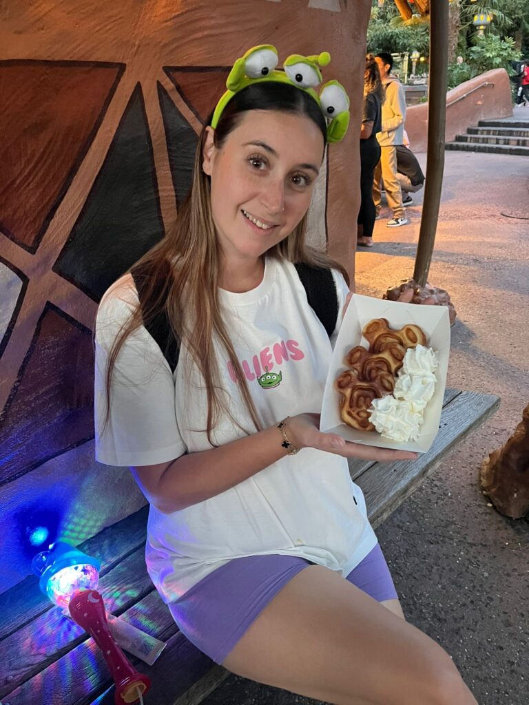 Rachel is sitting on a wooden bench, wearing a Toy Story alien headband and a white "Aliens" t-shirt. She is holding a tray of Mickey Mouse-shaped waffles with whipped cream. Next to her sits a glowing, colourful toy wand.