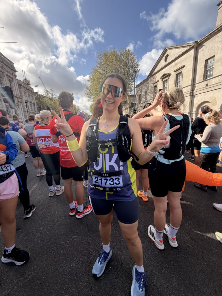 Rachel smiles and gives peace signs while standing in a crowd of runners. She wears a purple and yellow Guts UK running vest with race bib number 21733, reflective sunglasses, and a black hydration pack.