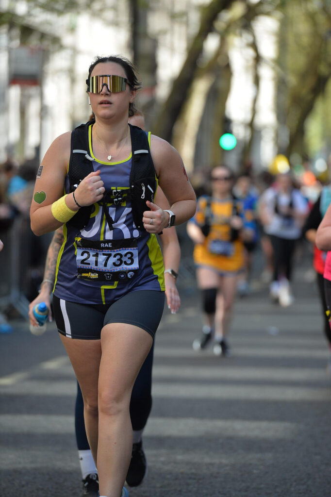 An action shot of Rachel running down a city street during the race. She is focused, wearing her Guts UK vest, hydration pack, and large reflective sunglasses.