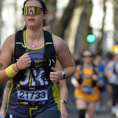 An action shot of Rachel running down a city street during the race. She is focused, wearing her Guts UK vest, hydration pack, and large reflective sunglasses.