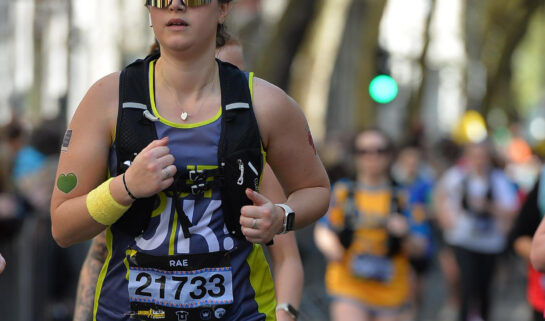An action shot of Rachel running down a city street during the race. She is focused, wearing her Guts UK vest, hydration pack, and large reflective sunglasses.