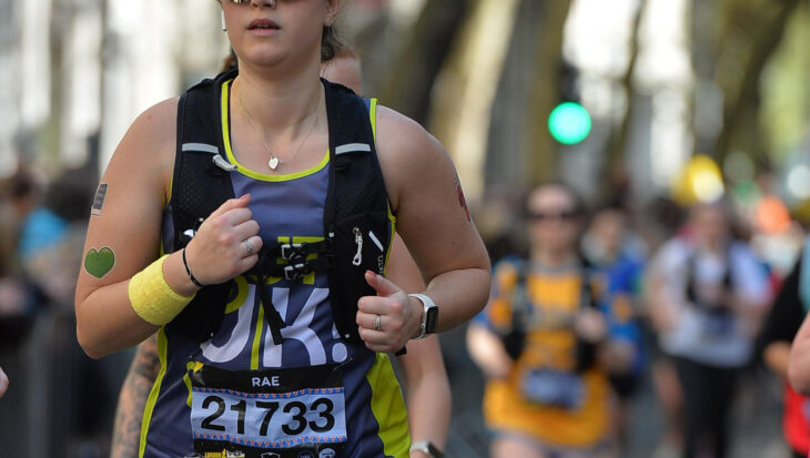 An action shot of Rachel running down a city street during the race. She is focused, wearing her Guts UK vest, hydration pack, and large reflective sunglasses.