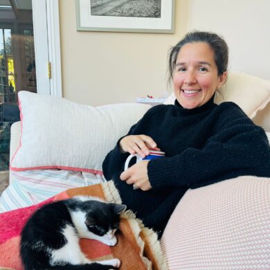 Sarah sits on a sofa with a cup of tea in her hand and a cat on her lap. She's wearing a turtle neck black jumper, and is smiling at the camera.