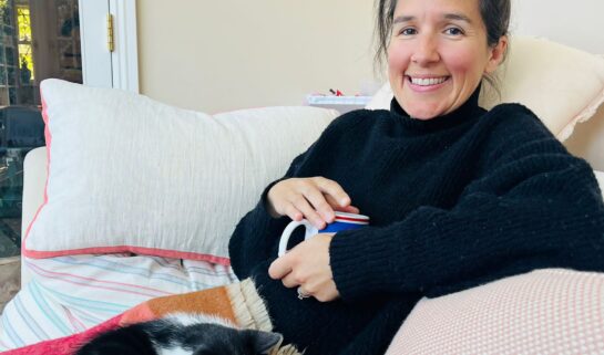Sarah sits on a sofa with a cup of tea in her hand and a cat on her lap. She's wearing a turtle neck black jumper, and is smiling at the camera.