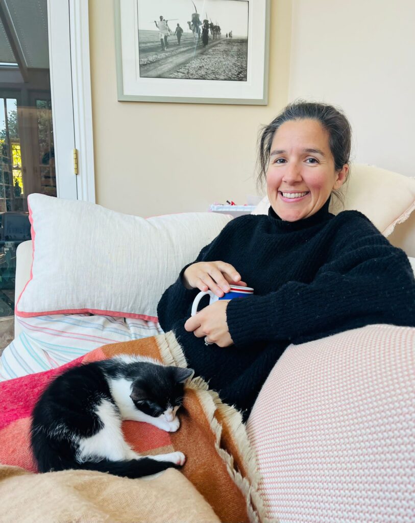 Sarah sits on a sofa with a cup of tea in her hand and a cat on her lap. She's wearing a turtle neck black jumper, and is smiling at the camera. 