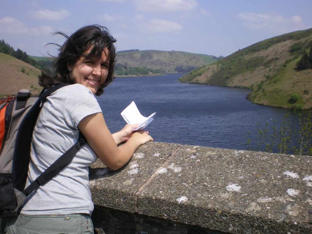 Sarah is looking at the camera over her shoulder and smiling. She is leaning on a wall with water and mountains in the background.