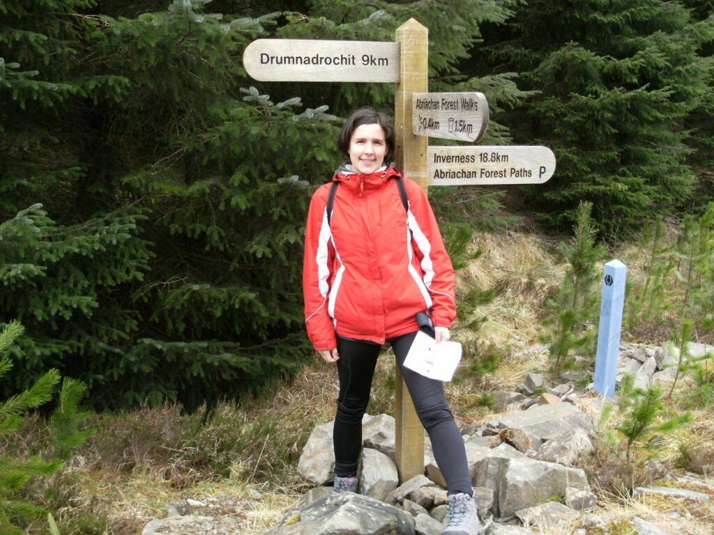 Sarah is wearing a red jacket, and walking boots and is stood in front of a directions sign, looking at the camera. 