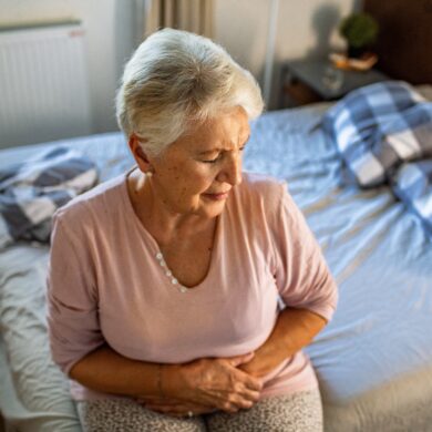 An older woman sits on the edge of her bed, holding her stomach in pain.