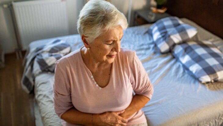An older woman sits on the edge of her bed, holding her stomach in pain.