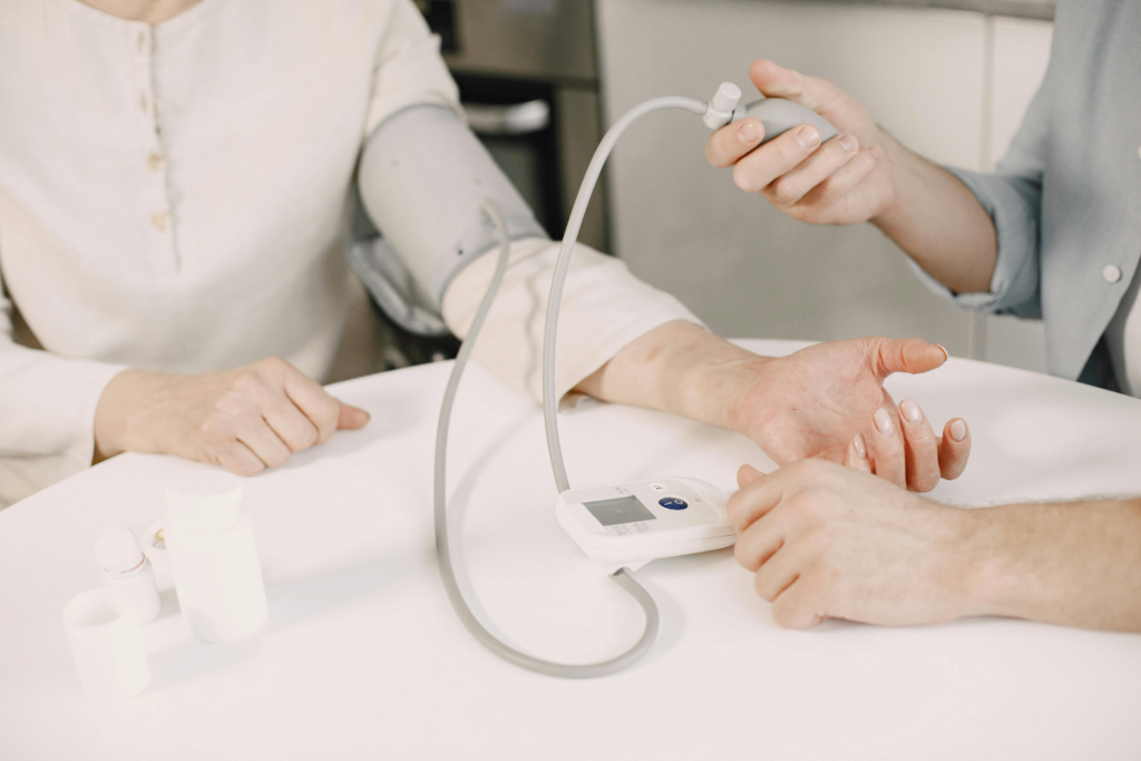 A close-up shot of a person having their blood pressure taken at a white table. An automated blood pressure cuff is wrapped around the upper arm of a person wearing a cream-colored shirt, while another person's hands are visible operating the manual pump and monitoring the digital display. Several small white medicine bottles sit on the table in the foreground.