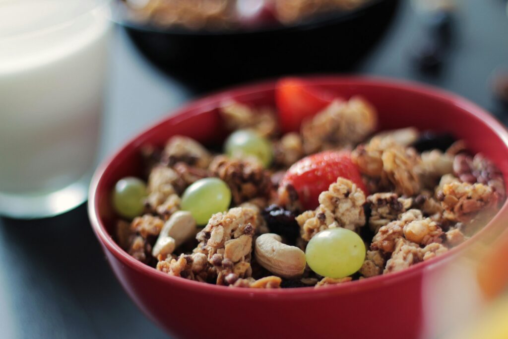 A close-up, slightly high-angle shot of a red bowl filled with granola, cashews, whole green grapes, and sliced strawberries. In the blurred background, a glass of milk and another dark bowl of granola sit on a dark surface.