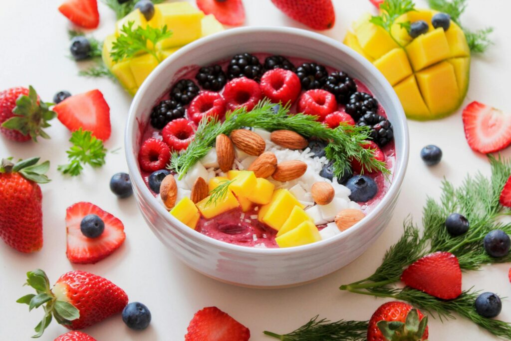 A top-down view of a vibrant pink smoothie bowl in a light gray ceramic bowl, set against a white background. The bowl is meticulously topped with rows of fresh raspberries, blackberries, blueberries, diced mango, whole almonds, and shredded coconut, garnished with a sprig of fresh dill. The surrounding table is artfully scattered with additional whole strawberries, blueberries, mango chunks, and sprigs of dill and parsley.