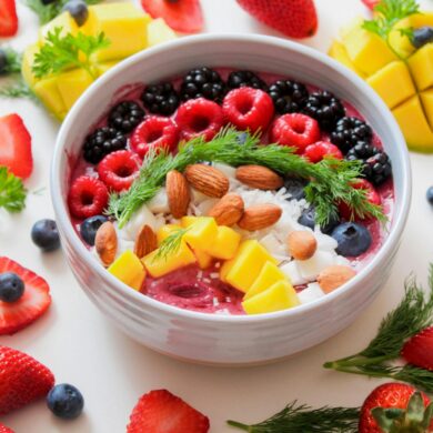 A top-down view of a vibrant pink smoothie bowl in a light gray ceramic bowl, set against a white background. The bowl is meticulously topped with rows of fresh raspberries, blackberries, blueberries, diced mango, whole almonds, and shredded coconut, garnished with a sprig of fresh dill. The surrounding table is artfully scattered with additional whole strawberries, blueberries, mango chunks, and sprigs of dill and parsley.