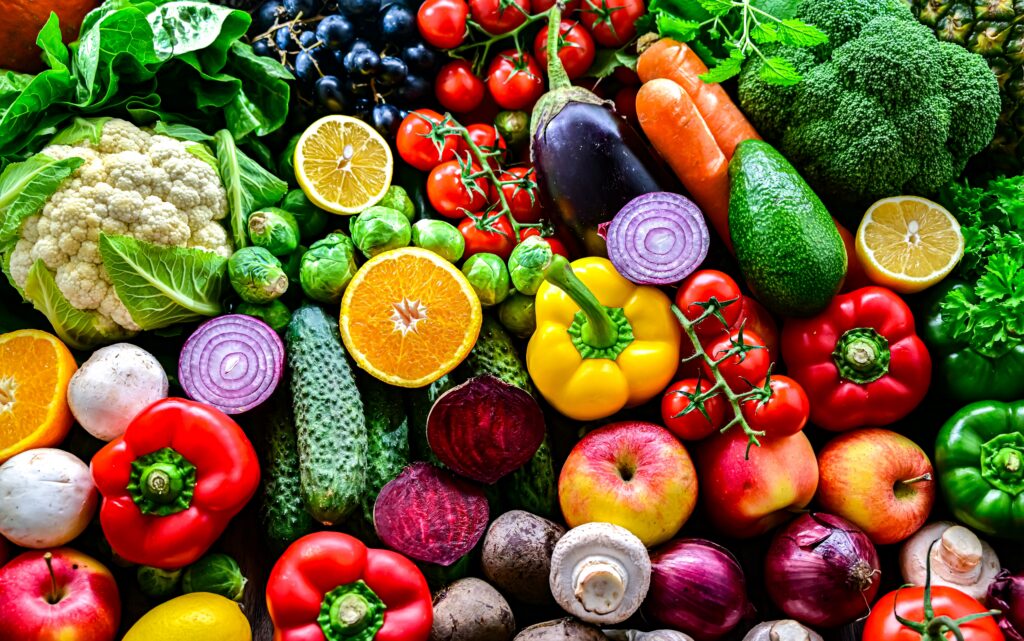 A vibrant, bird's-eye view of a dense variety of fresh fruits and vegetables, including peppers, broccoli, tomatoes, citrus, and leafy greens.