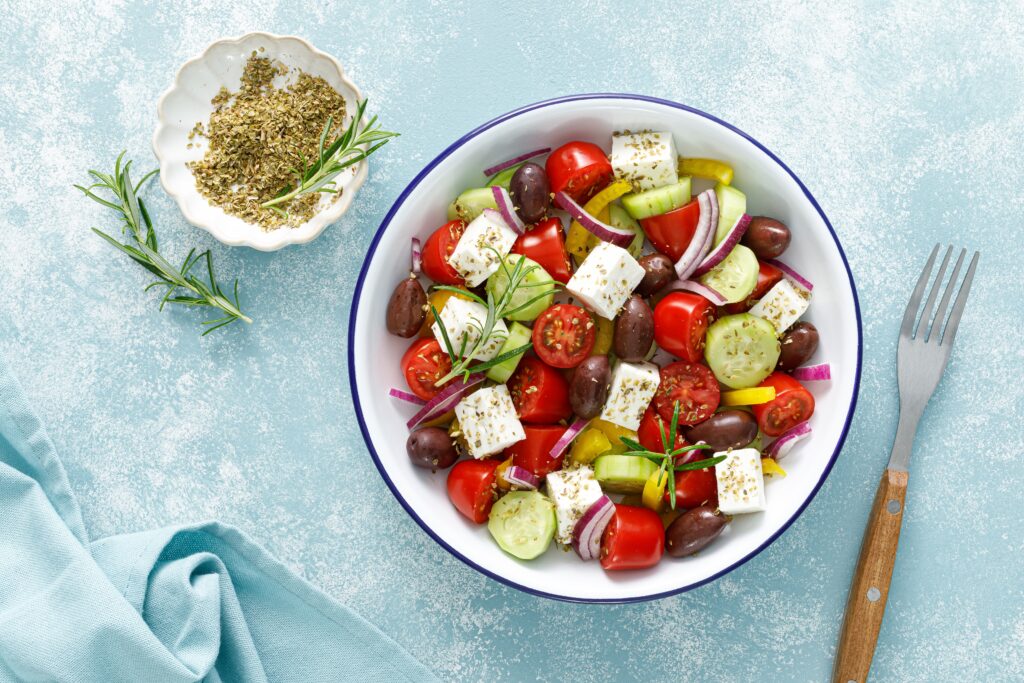 A top-down view of a fresh Greek salad in a white bowl, featuring tomatoes, cucumbers, feta cheese, olives, and red onions, seasoned with herbs and served on a light blue textured surface.