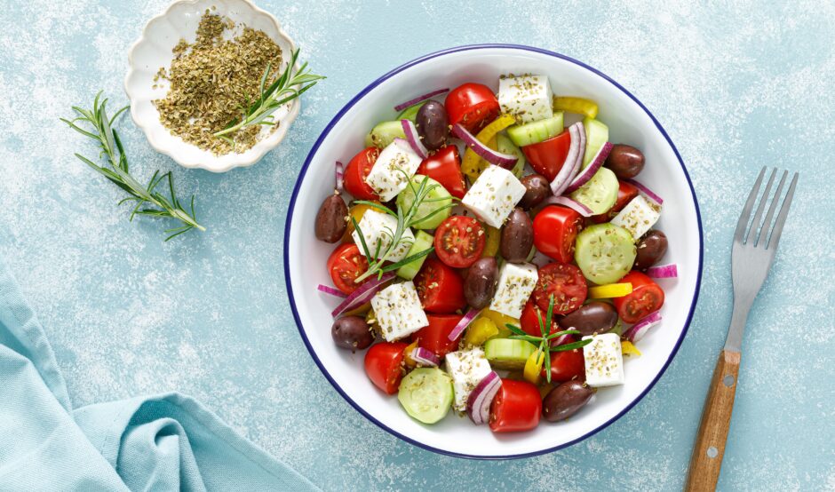 A top-down view of a fresh Greek salad in a white bowl, featuring tomatoes, cucumbers, feta cheese, olives, and red onions, seasoned with herbs and served on a light blue textured surface.