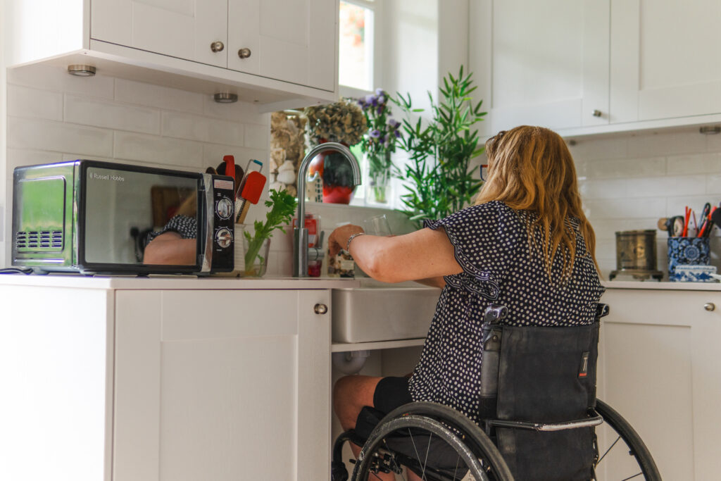 A woman with long reddish-blonde hair sits in a black manual wheelchair, facing a white farmhouse-style sink. The kitchen features white cabinetry and subway tiles, with a black Russell Hobbs microwave on the counter to her left.