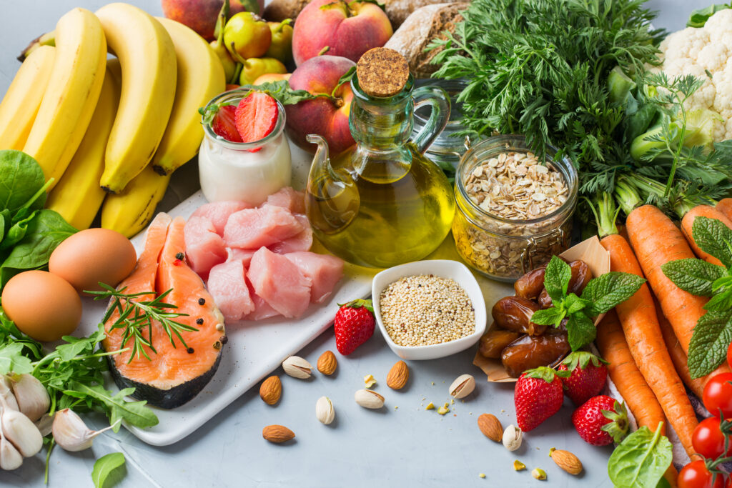 A colourful flat lay arrangement of Mediterranean diet foods including fresh salmon, cubed poultry, eggs, olive oil, Greek yogurt with strawberries, bananas, peaches, carrots, tomatoes, nuts, seeds, and leafy greens.