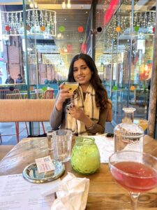 Sehba is sat down at a restaurant with a table in front of her and smiling as she holds up a yellow cocktail in a coupe glass. She is wearing a grey long-sleeved top and a cream and black checked scarf. There are several drinks on the table.