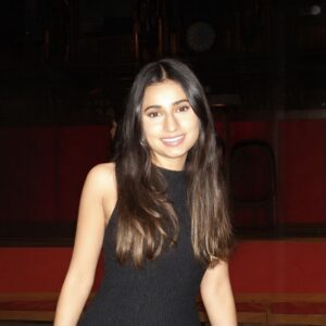 Sehba has long dark hair and is smiling warmly at the camera. She is wearing a sleeveless black dress. The background is dimly lit with dark wood panelling and a hint of red tiered seating.