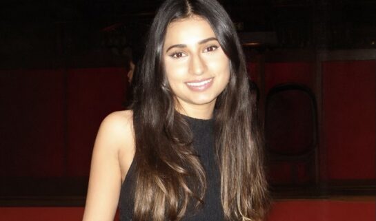 Sehba has long dark hair and is smiling warmly at the camera. She is wearing a sleeveless black dress. The background is dimly lit with dark wood panelling and a hint of red tiered seating.