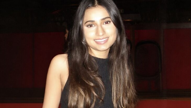 Sehba has long dark hair and is smiling warmly at the camera. She is wearing a sleeveless black dress. The background is dimly lit with dark wood panelling and a hint of red tiered seating.