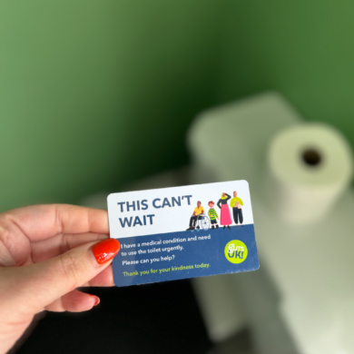 A person is holding a Can't Wait Card in their hand. Behind the card is a toilet with a roll of toilet roll on top.