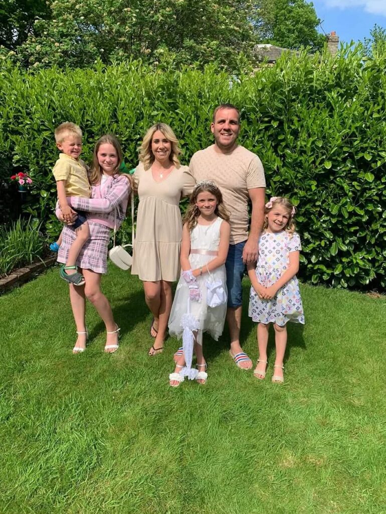 A family group photo taken outdoors on a sunny day in front of a tall green hedge. A man and a woman stand in the centre, surrounded by four children. On the left, an older girl in a pink plaid outfit holds a young boy in a yellow shirt. In the centre, a young girl wears a white dress with a tiara and holds a white lace umbrella, and to the right, another young girl smiles in a patterned white dress.