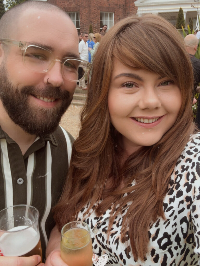 A close-up selfie of a smiling man and woman holding drinks at an outdoor party. The man has a beard, clear-rimmed glasses, and a striped polo shirt. The woman has long brown hair with bangs and is wearing a leopard print top. Other guests and a brick building are visible in the blurred background.