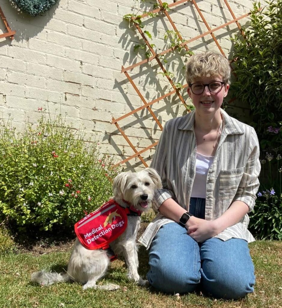 Leah and her medical alert dog, Doris, sitting together outside on the grass in front of a white brick wall with a wooden trellis. Leah has short blonde hair and glasses and is wearing a striped shirt and jeans. Doris, a small white, scruffy dog, sits upright next to her wearing a bright red vest that reads "Medical Detection Dogs."