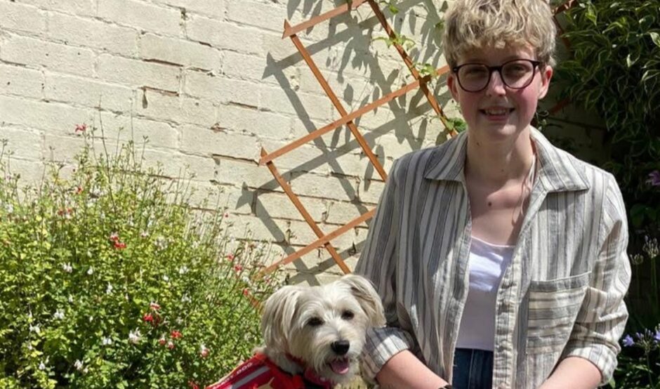 Leah and her medical alert dog, Doris, sitting together outside on the grass in front of a white brick wall with a wooden trellis. Leah has short blonde hair and glasses and is wearing a striped shirt and jeans. Doris, a small white, scruffy dog, sits upright next to her wearing a bright red vest that reads "Medical Detection Dogs."
