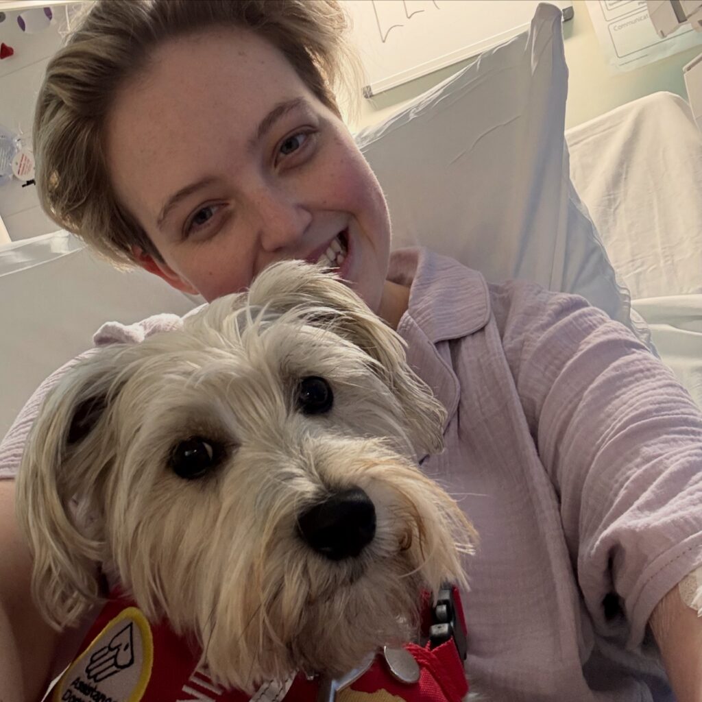 A selfie of Leah smiling from a hospital bed, leaning in close beside their medical alert dog, Doris. Leah has short light hair and is wearing purple pyjamas. Doris, her small scruffy white assistance dog, looks directly at the camera while wearing her red assistance dog vest.