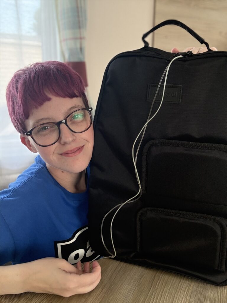 A close-up shot of Leah smiling as they lean next to a large black medical backpack resting on a table. Clear tubing runs from the top zipper of the backpack. Leah has short purple hair and glasses and is wearing a blue sweatshirt.