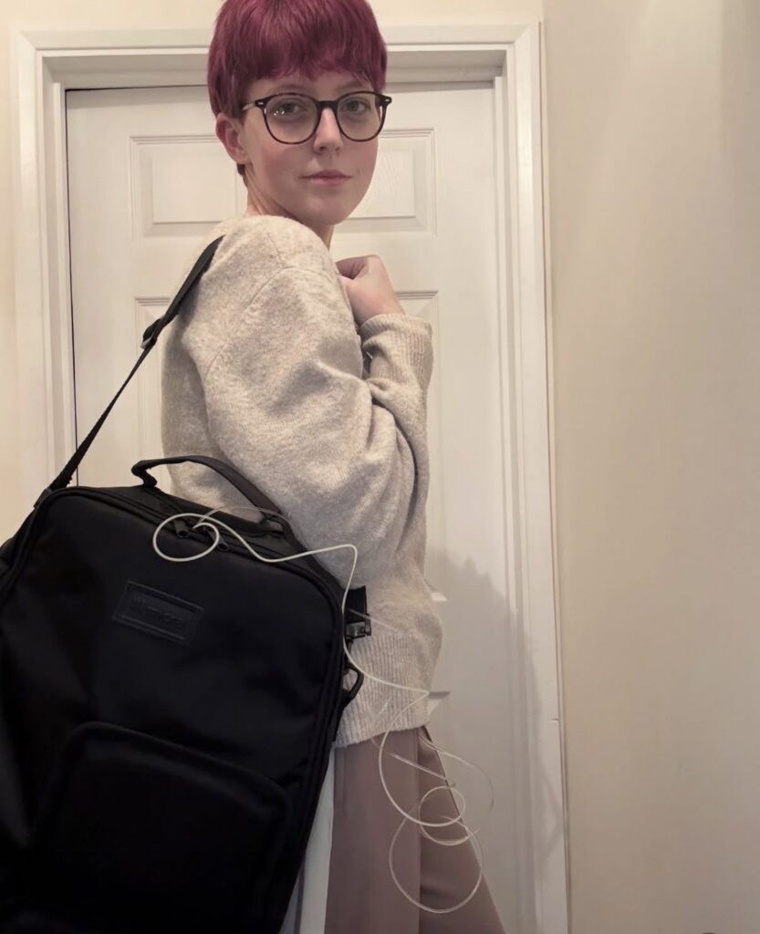 A side-view portrait of Leah, a person with short purple hair and glasses, wearing a beige knitted sweater. They are looking at the camera while wearing a black backpack on one shoulder. Clear medical tubing extends from inside the backpack and loops down past their side.