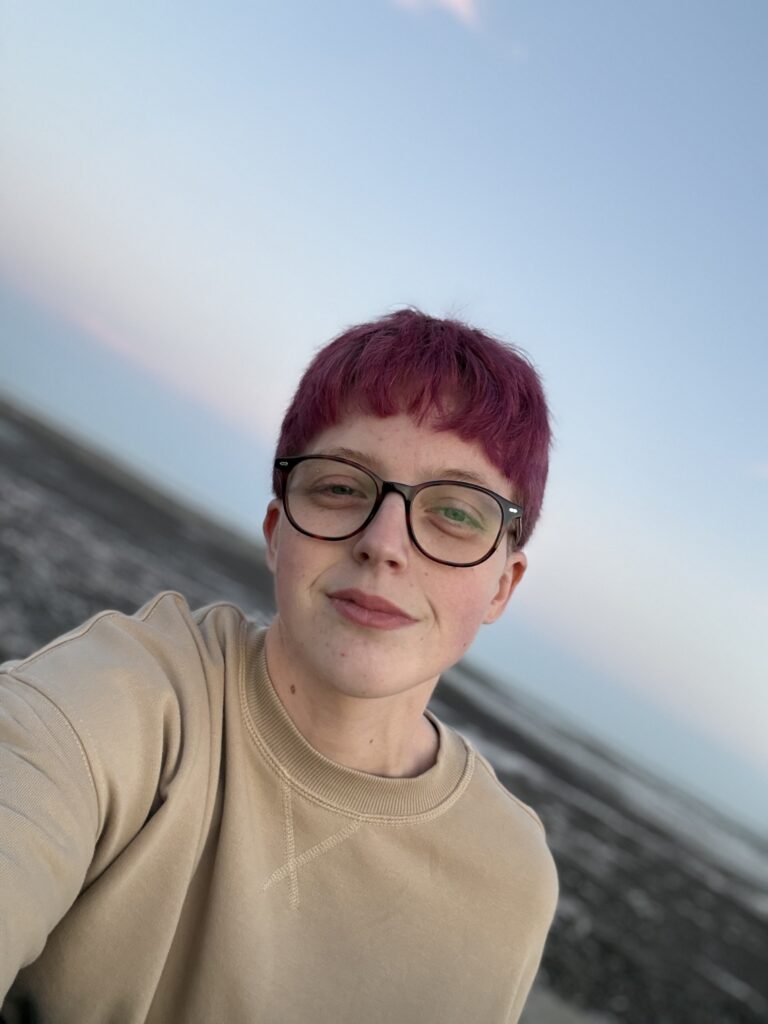 A close-up outdoor selfie of Leah, featuring their short purple hair and dark-rimmed glasses. They are wearing a tan sweatshirt, looking into the camera with a neutral expression. The background shows a slanted view of a beach landscape under a clear sky.