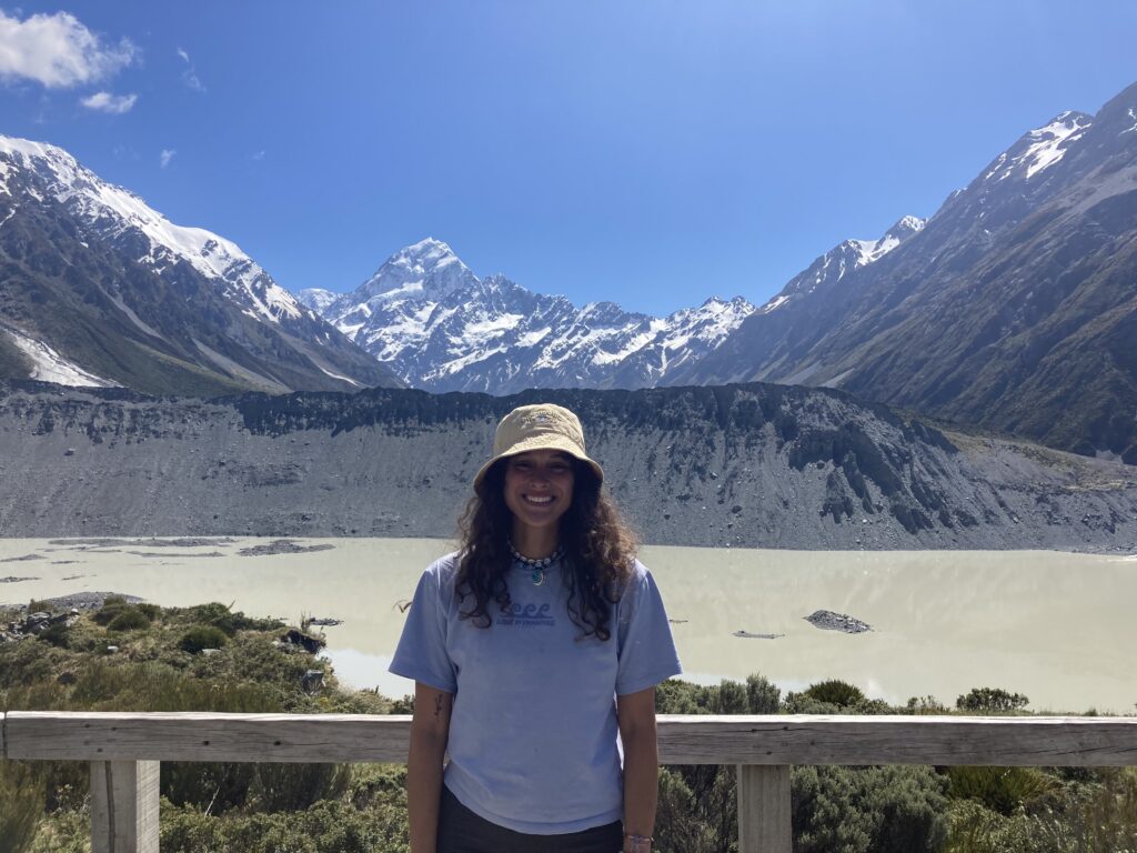 A medium shot of a smiling woman with long curly brown hair, wearing a beige bucket hat, a light blue t-shirt, and a beaded necklace. She is standing behind a rustic wooden railing. The background features a vast, light-colored glacial lake, a large gray moraine wall, and a striking snow-capped mountain peak under a clear blue sky.