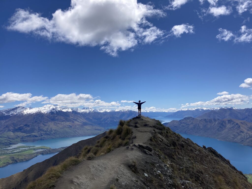 A wide, scenic landscape shot of a person standing at the very peak of a steep, rocky mountain ridge with their arms wide open. The background showcases a breathtaking panoramic view of deep blue lakes winding through rolling green hills and a massive range of snow-capped mountains under a bright blue sky with scattered white clouds.