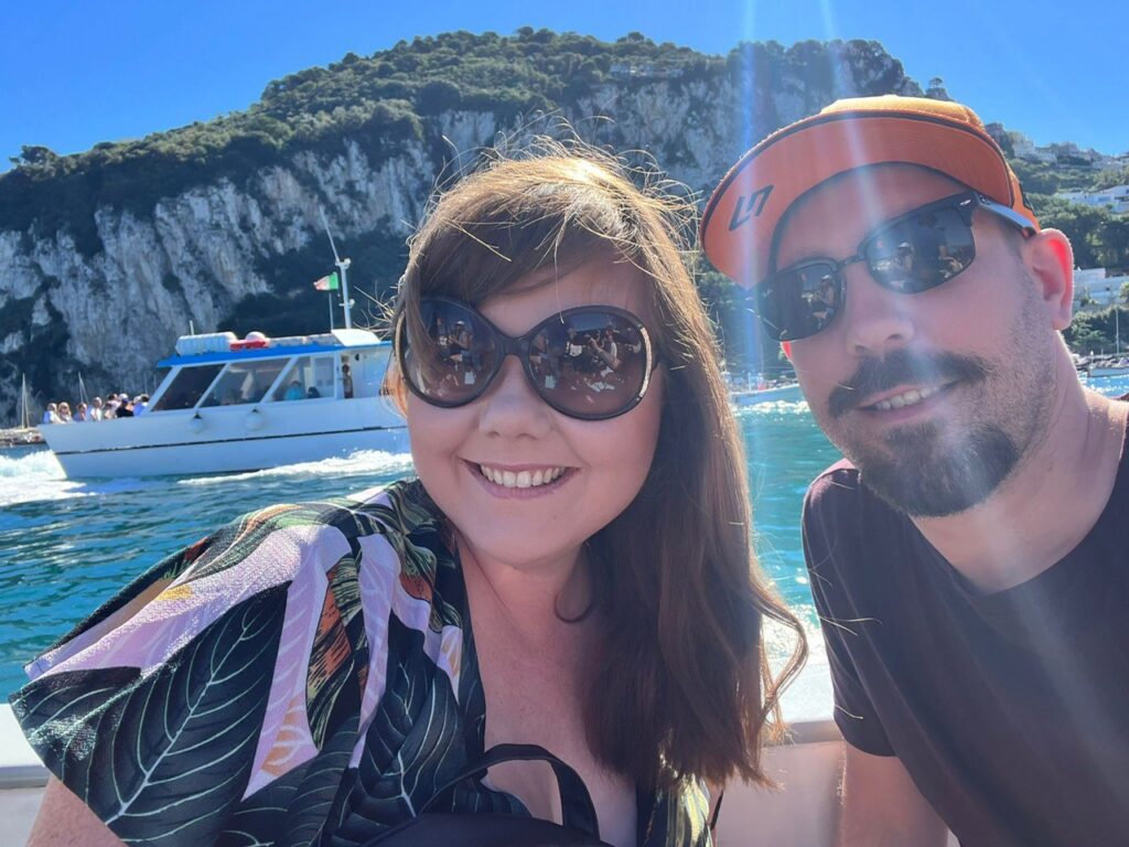 A sunny selfie of a smiling woman in oversized round sunglasses and a bearded man wearing a bright orange cap and sunglasses. They are on a boat with turquoise blue water and a massive rocky cliff in the background.