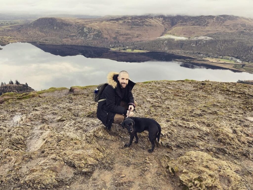 A man in a winter coat with a faux-fur hood crouches next to a black dog on a rocky, elevated peak. A large, calm lake reflecting a cloudy sky and rolling mountains stretches out below them.
