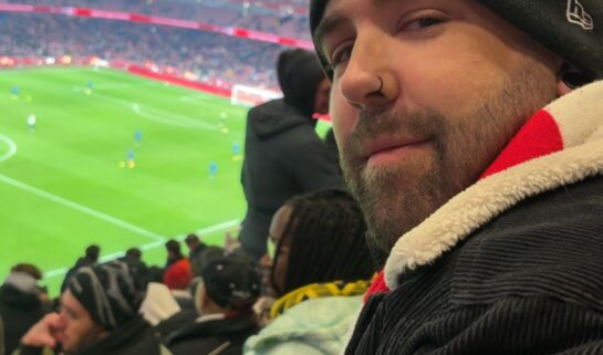A side-profile selfie of a bearded man with a nose ring wearing a New York Yankees beanie and a black corduroy jacket. He is sitting in red stadium seats overlooking a brightly lit green football pitch during a match.