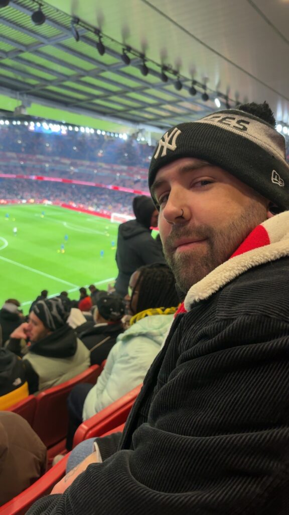 A side-profile selfie of a bearded man with a nose ring wearing a New York Yankees beanie and a black corduroy jacket. He is sitting in red stadium seats overlooking a brightly lit green football pitch during a match.