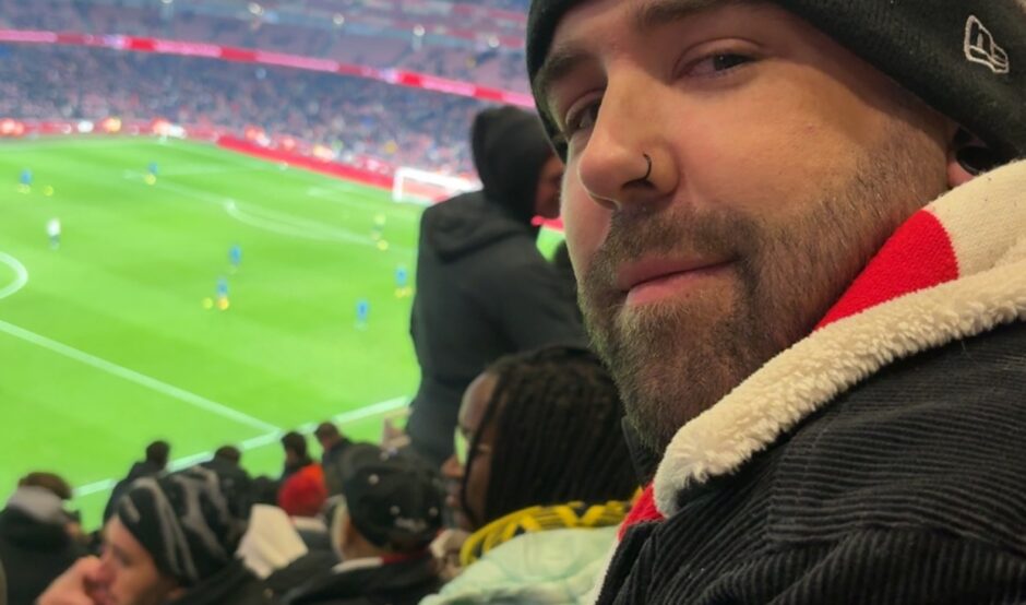 A side-profile selfie of a bearded man with a nose ring wearing a New York Yankees beanie and a black corduroy jacket. He is sitting in red stadium seats overlooking a brightly lit green football pitch during a match.