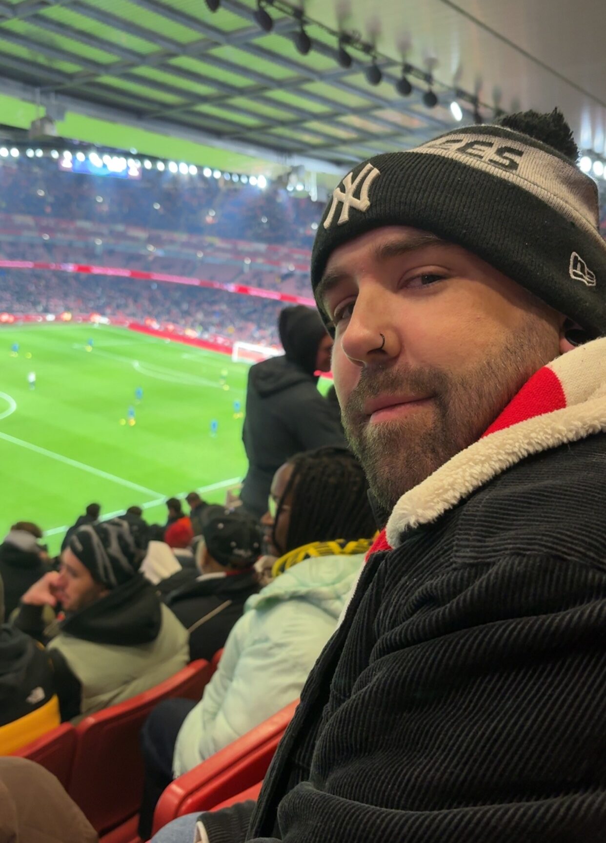 A side-profile selfie of a bearded man with a nose ring wearing a New York Yankees beanie and a black corduroy jacket. He is sitting in red stadium seats overlooking a brightly lit green football pitch during a match.