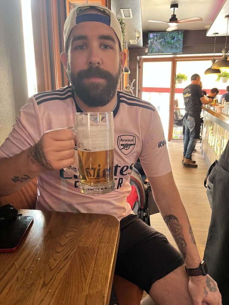 A bearded man wearing a pink Arsenal football jersey and a backwards baseball cap sits at a wooden table in a bar, holding up a large glass mug of beer. His arms have visible tattoos, and the bar's interior is visible in the background.