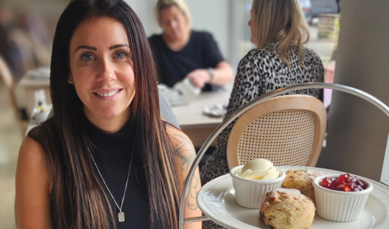 A woman named Rebecca with long dark hair, wearing a black sleeveless top and a silver necklace, smiling at the camera. She is seated at a restaurant table enjoying afternoon tea. In front of her is a three-tiered cake stand filled with scones, clotted cream, jam, pastries, and finger sandwiches, alongside a white teapot, teacup, and various table settings.