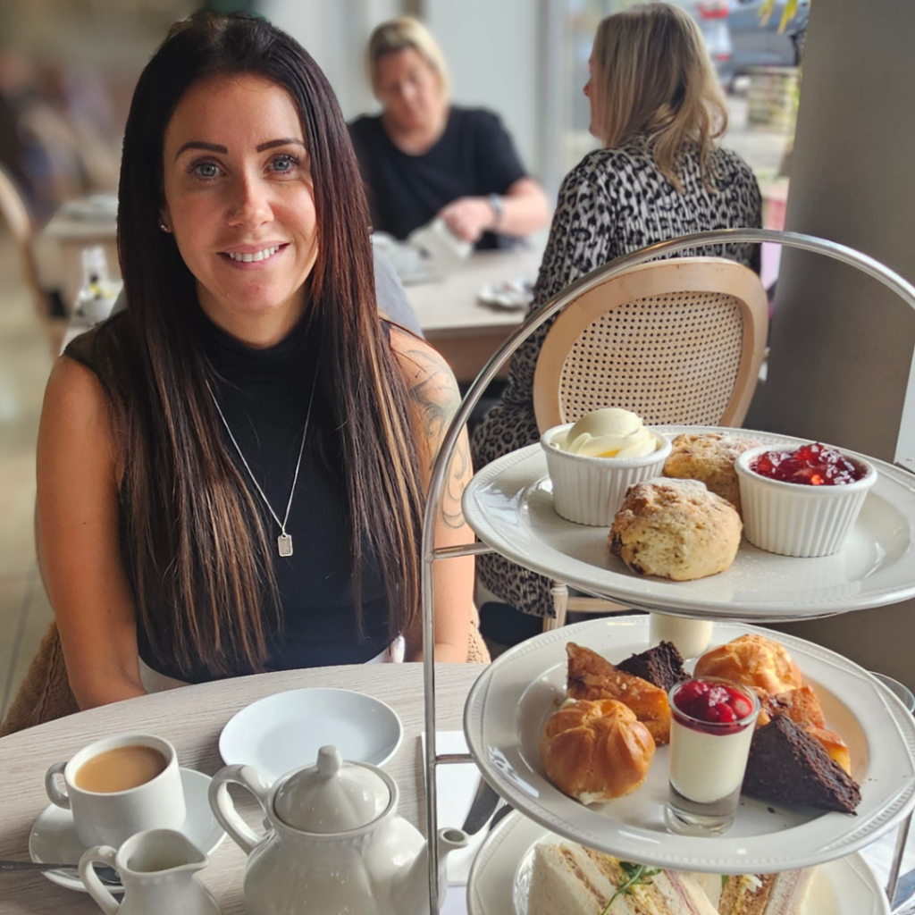 A woman named Rebecca with long dark hair, wearing a black sleeveless top and a silver necklace, smiling at the camera. She is seated at a restaurant table enjoying afternoon tea. In front of her is a three-tiered cake stand filled with scones, clotted cream, jam, pastries, and finger sandwiches, alongside a white teapot, teacup, and various table settings.