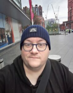 A close-up selfie of a man wearing a navy blue beanie and glasses on an outdoor pavement