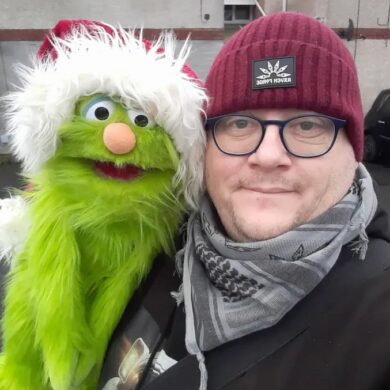 A selfie of a man wearing a maroon beanie and glasses, smiling next to a green Kermit-like puppet wearing a Santa hat.