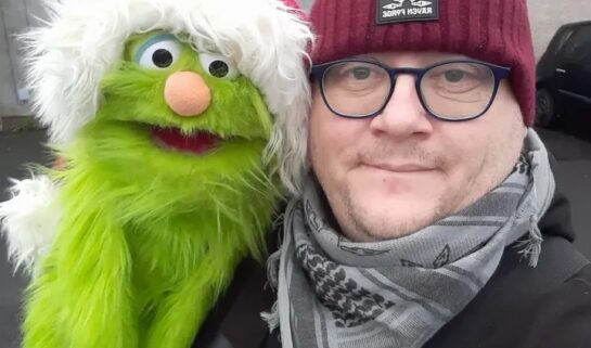 A selfie of a man wearing a maroon beanie and glasses, smiling next to a green Kermit-like puppet wearing a Santa hat.